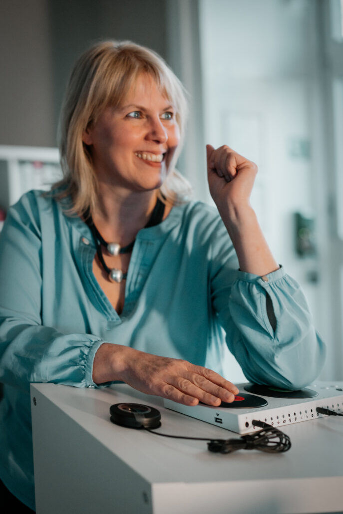 Susanne Baumer Leiterin Test.Labor Barrierefreiheit mit einem freundlichen Lächeln und der Hand auf einem Gerät am Schreibtisch.