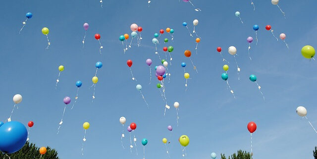 Viele bunte Luftballons steigen in den blauen Himmel. Darunter Baumwipfel. Ballons haben einen Zettel an der Schnur.